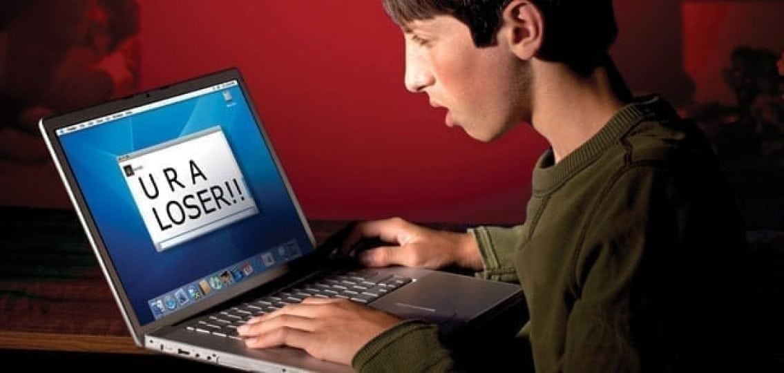 A boy sitting in front of his laptop looking at the screen with the words reading you are a loser. comforted by friend with bullies in the back